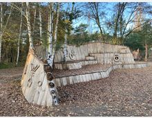 Holzstruktur in einem Wald, gestaltet wie eine abstrakte Skulptur mit geometrischen Formen und Mustern. Im Hintergrund stehen Birken und andere Bäume auf einem mit Laub bedeckten Boden.