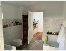 Das Bild zeigt ein Badezimmer mit beigefarbenen Wandfliesen und einem Holzboden. Auf der linken Seite befindet sich eine wandmontierte Toilette, darüber Ablageflächen mit verschiedenen Gegenständen. Rechts ist eine Badewanne zu sehen, entlang deren Rand Spielzeug und andere Objekte platziert sind. Eine geöffnete Tür im Hintergrund führt in einen Raum mit Holzboden und Pflanzen auf einem Sideboard.