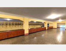 Großer Garderobenbereich mit langen Holztresen, gelben Wänden und zahlreichen Metall-Kleiderhaken an der Wand. Der Boden besteht aus grauen Fliesen, und die Beleuchtung erfolgt über mehrere helle Deckenleuchten.