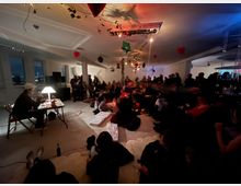 Ein dimm beleuchteter Raum mit weißen Wänden, dekoriert mit hängenden Herzen, Luftballons und Lichterketten. Im Vordergrund sitzt eine Person an einem Tisch mit Lampe, während im Hintergrund eine Gruppe von Menschen im Publikum auf Stühlen und auf dem Boden sitzt.