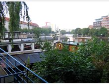 Ufer eines Flusses mit mehreren angedockten Booten, umgeben von dichtem Grün. Im Hintergrund sind Gebäude mit roten und modernen Fassaden sowie Baumreihen entlang des Wassers zu sehen.