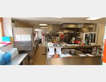 Industrieküche mit Edelstahl-Arbeitsflächen, großen Küchengeräten wie Herden und Öfen sowie Regalen mit Küchenutensilien. Ein Mitarbeiter arbeitet im Hintergrund, und auf den Arbeitsflächen liegen Papiere, Gewürze und Flaschen.