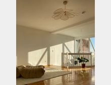 Heller Wohnraum mit minimalistischem Design, Holzboden und weißen Wänden. Ein großes Fenster mit Balkonelementen lässt Tageslicht herein, vor dem Geländer steht eine grüne Zimmerpflanze, und auf dem Teppich liegen ein beiger Sessel und ein passender Hocker.