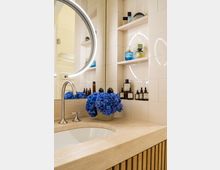 Modernes Badezimmer mit einer Waschtischplatte aus beigem Stein, einem eingelassenen Waschbecken und einem runden beleuchteten Spiegel. Im Hintergrund befinden sich weiße Fliesen und eingebauten Regale mit Pflegeprodukten; ein Strauß blauer Hortensien steht auf der Ablage.