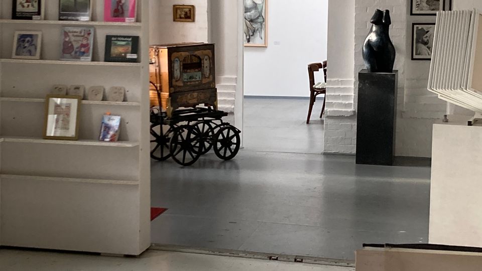 Innenraum einer Kunstgalerie mit weißen Ziegelwänden und grauem Boden, Blick durch einen breiten Rundbogen in einen weiteren Ausstellungsraum. Links stehen Regalflächen mit Büchern und Druckgrafiken, rechts sind gerahmte Bilder an der Wand und eine dunkle Skulptur auf einem Sockel zu sehen.