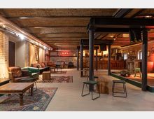 Gemütlich eingerichteter Veranstaltungsraum mit Backsteinwänden und freiliegenden Deckenbalken. Der Raum verfügt über eine Bar im Hintergrund, bunte Teppiche, Polstermöbel sowie eine kleine Bühne mit einem Mikrofon und Verstärker.