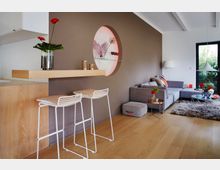 Modernes Wohnzimmer mit offener Küche, hellbraunem Holzfußboden und einer grauen L-förmigen Couch. Eine Holztheke mit zwei weißen Barhockern grenzt an eine Wand mit einem runden Regal, während große Fenster Licht auf einen Teppich und Couchtisch werfen.