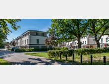 Moderne Reihenhaussiedlung mit hellen Fassaden und großen Fenstern, umgeben von Grünflächen, Bäumen und einem gepflasterten Weg im Vordergrund. Im Hintergrund sind weitere ähnliche Gebäude sichtbar.