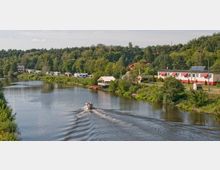 Flusslandschaft mit einem Boot auf dem Wasser, umgeben von Bäumen und Campingfahrzeugen am Ufer. Im Hintergrund befinden sich Gebäude, darunter ein flaches Gebäude mit roten Markisen und Solaranlagen auf dem Dach.
