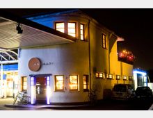 Ein zweistöckiges, gelblich beleuchtetes Gebäude mit großen, rechteckigen Fenstern bei Nacht. Ein beleuchtetes Schild mit der Aufschrift „Anhalt“ und ein Eingang mit Lichtröhren befinden sich im Vordergrund, links daneben ein überdachter Bereich mit Glasdach.