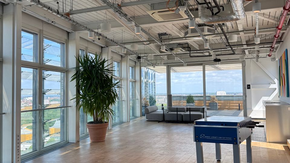 Open-plan modern office lounge with floor-to-ceiling windows, wood flooring, and an exposed industrial ceiling with ducts, pipes, and hanging lights. A foosball table, large potted plant, and seating area sit near glass doors leading to a terrace with a city view.