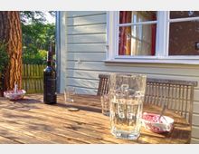 Ein Holztisch mit Getränkegläsern, einer Wasserkanne und einer Weinflasche steht vor einer holzverkleideten Hausfassade mit weißem Fensterrahmen. Im Hintergrund sind ein Baum und ein Zaun in einem grünen Garten sichtbar.