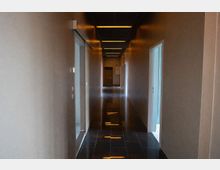 Langer, schmaler Innenflur in einem modernen Gebäude mit glatten grauen Wänden, mehreren Türöffnungen auf beiden Seiten und dunklem, glänzend gefliestem Boden. An der Decke verlaufen lineare Einbauleuchten, die sich im Boden spiegeln, am Ende des Flurs ist eine geschlossene Tür zu sehen.