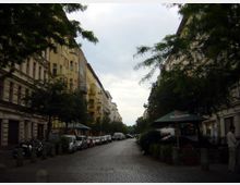 Straßenszene in einem Wohnviertel mit mehrstöckigen Altbauten auf beiden Seiten, die von Bäumen gesäumt werden. Am rechten Bildrand befinden sich ein Café mit einem grünen Sonnenschirm und parkende Autos entlang der Straße.