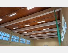 Blick in eine Sporthalle mit hoher Holzdecke, weißen Querträgern und langen Deckenleuchten. An den Wänden befinden sich große Fenster sowie ein gespanntes Schutznetz, daneben hängt ein grünes Seitennetz.