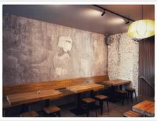 Ein modernes Café mit schlichten Holztischen, passenden Holzhockern und einer langen Sitzbank entlang der Wand. Die Wand ist mit einer Beton-Optik gestaltet, die ein gemaltes Motiv einer Frau mit Fächer zeigt, daneben eine teilweise freigelegte weiße Backsteinwand; eine einzelne Hängelampe beleuchtet den Raum.