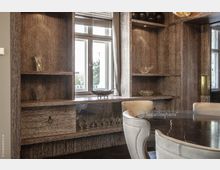 Holzvertäfelter Innenraum mit eingebautem Regal, in dem Glasdekorationen ausgestellt sind, und einem Fenster, durch das Tageslicht einfällt. Im Vordergrund steht ein runder Tisch mit einer dunklen, marmorierten Oberfläche, umgeben von hell gepolsterten Stühlen.