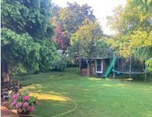 Garten mit großer Rasenfläche, mehreren Bäumen und Sträuchern. Im Hintergrund befinden sich ein Holzspielhaus mit grüner Rutsche und ein Trampolin, im Vordergrund stehen Blumentöpfe mit pinkfarbenen Hortensien und ein Wasserschlauch.