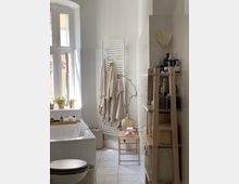Bright white-tiled bathroom with a bathtub under a tall window, small potted plants on the sill, and a towel radiator holding hanging robes. A wooden ladder-style shelf with toiletries and baskets stands beside a small stool and decorative vase on the tiled floor.