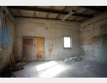 Innenraum einer unfertigen Werkstatt oder Garage mit rohen Beton- und verputzten Wänden, freiliegender Decke mit Holzbalken und einer hängenden Leuchtstofflampe. An der Rückwand stehen eine provisorisch verkleidete Tür und ein kleines Fenster; auf dem Betonboden liegen einige Bausäcke und Baumaterialien.