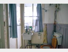 Kleines Badezimmer mit einfacher Ausstattung, bestehend aus einem Waschbecken, einer Toilette mit Holzsitz und einigen sichtbaren Rohren und Leitungen. Im Hintergrund befindet sich eine verglaste Tür mit blauen Vorhängen, die teilweise Tageslicht hereinlässt.