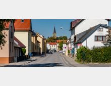 Straßenszene in einem Wohngebiet mit zweistöckigen Häusern und roten Dächern, links und rechts der Straße. Im Hintergrund ist eine Kirche mit einem hohen Turm und schwarzer Spitze zu sehen, umgeben von Bäumen.