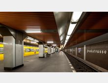U-Bahn-Station mit dem Namen „Pankstraße“; zu sehen sind ein gelber U-Bahn-Zug auf der linken Seite, braune Wandverkleidungen mit Stationsnamen, eine dunkle Gleisanlage und ein mit Metallstützen und Holzdecke versehener Wartebereich. Die Beleuchtung besteht aus rechteckigen Leuchtstofflampen.