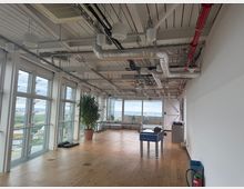 Open-plan modern office lounge with a wooden floor, floor-to-ceiling windows overlooking a city, and an exposed industrial ceiling with ducts and pipes. The space includes a potted plant, a foosball table, and a sofa seating area near large windows.