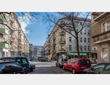 Straßenszene in einem Wohngebiet mit mehrstöckigen Altbauten, die Balkone und große Fenster haben. Die Straße ist mit Kopfsteinpflaster gepflastert, gesäumt von geparkten Autos und einigen kahlen Bäumen, unter einem klaren blauen Himmel.