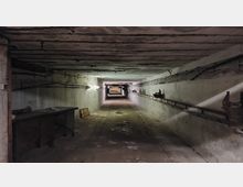Ein langer, leerer unterirdischer Tunnel mit Betonwänden und einer flachen Decke. An den Wänden verlaufen Rohre und Kabel, während an der linken Seite ein alter Tisch und weitere Gegenstände stehen.