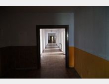 Ein langer, leerer Korridor mit Holzboden und weißen Wänden, teilweise mit gelber Farbe und braunen Fliesen verkleidet. Der Flur ist durch Wandöffnungen unterteilt, hat links Fenster und Heizkörper sowie am Ende eine Tür mit Fluchtweg-Schild.