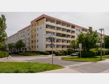 Wohngebäude in einer Wohnstraße mit fünf Etagen und Balkonen, die in Pastelltönen gestrichen sind. Im Vordergrund befinden sich Verkehrszeichen, gepflasterte Gehwege, Grünflächen und Bäume.