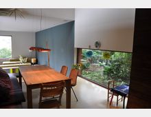 Moderne Wohnküche mit großem Holztisch, mehreren Stühlen und einer hängenden Lampe. Ein großes Fenster bietet Blick in einen grünen Garten, während eine blaue Wand und helle Möbel den Raum definieren.