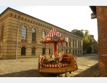 Historisches Backsteingebäude mit vielen Rundbogenfenstern auf einem gepflasterten Platz; im Vordergrund ein kleines nostalgisches Kinderkarussell mit rot-weiß gestreiftem Dach und Pferdefiguren. Im Hintergrund sind Bäume und ein weiteres Gebäude zu sehen.