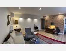 Wohnraum mit gemischtem Stil: Eine helle Wand mit integrierter Beleuchtung grenzt an eine freiliegende Ziegelwand. Im Raum befinden sich ein grauer Sessel, eine beige Couch, eine antike Kommode, dekorative Lampen und ein roter Teppich auf einem Holzboden.