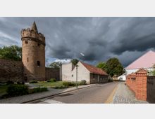 Eine historische Altstadtstraße mit einem aus Backstein gebauten, runden Wehrturm, flankiert von einer Steinmauer links. Rechts befinden sich kleine Häuser mit roten Ziegeldächern, und im Hintergrund ziehen dunkle Gewitterwolken am Himmel auf.