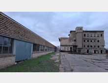 Industriegelände mit verwitterten Gebäuden aus Ziegelstein und Beton. Ein langes flaches Gebäude mit großen Fenstern steht links, während rechts ein mehrstöckiges, stark gealtertes Gebäude mit zerbrochenen Fenstern und abgeschlagenen Türen zu sehen ist; der Boden besteht aus Betonplatten mit vereinzeltem Grasbewuchs.