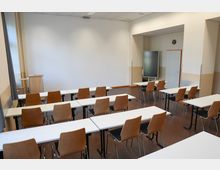 Ein leerer Seminarraum mit weißen Tischen und Holzstühlen, angeordnet in Reihen. Im Hintergrund befinden sich ein Rednerpult, eine Tür, eine Wanduhr und ein digitales Whiteboard.