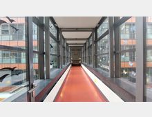 Ein verglaster Übergang mit einer roten Bodenfläche, flankiert von schwarzen Metallrahmen und Fenstern, die mit Aufklebern von Vögeln und Schmetterlingen dekoriert sind. Im Hintergrund ist ein modernes Gebäude mit orangefarbenen und grauen Fassadenelementen sichtbar.