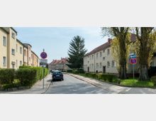 Wohnstraße in einem Wohngebiet mit zweigeschossigen Reihenhäusern, umgeben von gepflegten Hecken und Bäumen. Ein Gehweg verläuft entlang der Straße, auf der parkende Autos und Verkehrsschilder zu sehen sind.
