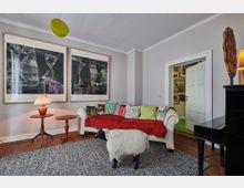 Wohnzimmer mit hellgrauen Wänden, einem cremefarbenen Sofa mit bunten Kissen und rotem Überwurf, zwei großen abstrakten Kunstwerken an der Wand und einem Teppich in Schwarz-Weiß. Im Vordergrund steht eine dekorative Schafskulptur, und im Raum befinden sich ein Klavier, zwei Beistelltische und eine Lampe.