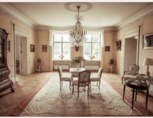 Eleganter Wohnraum mit klassischen Möbeln aus dem 18. oder 19. Jahrhundert. Der Raum hat einen großen Kronleuchter, einen Holzboden mit einem dekorativen Teppich, eine Sitzgruppe im Zentrum, großzügige Fenster mit weißen Vorhängen und Wände in Pastellfarben, gesäumt von Gemälden und Ornamenten.