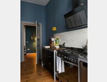 Moderne Küche mit dunkelblauen Wänden, Holzboden und einer Edelstahlküchenzeile mit großem Gasherd. Im Vordergrund steht eine Arbeitsplatte mit Tulpen, im Hintergrund führt eine geöffnete Tür in einen weiteren Raum mit gedämpfter Beleuchtung.