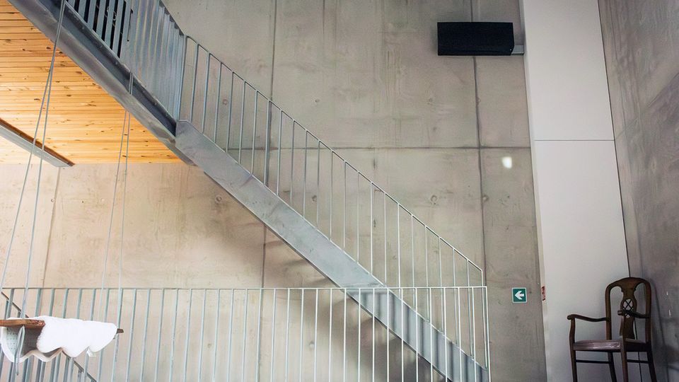 Innenraum mit hohen Sichtbetonwänden und einer geraden Metalltreppe mit Geländer, die diagonal zu einer oberen Ebene führt. Links ist eine Holzdecke zu sehen; rechts hängen ein Bildschirm und ein Ausgangsschild, daneben steht ein einzelner Holzstuhl.