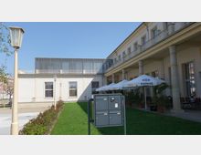 Außenbereich eines Gebäudes mit cremefarbener Fassade, hohen Fenstern und einer Säulenreihe, die einen überdachten Gang stützen. Im Vordergrund ein kleiner grüner Rasen mit einer Briefkastenanlage, rechts Sonnenschirme und Sitzplätze einer Außengastronomie.