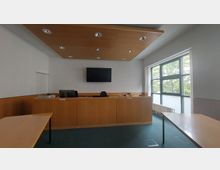 Ein moderner Gerichtssaal mit Holzeinrichtung, bestehend aus einem Richterpult mit einem Bildschirm an der Wand dahinter und großen Fenstern rechts, die Tageslicht hereinlassen. Der Raum ist mit grünem Teppichboden ausgelegt, und an den Seiten stehen schlichte Holztische.