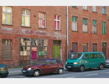 Backsteinfassade eines älteren Wohn- oder Geschäftshauses mit teilweise verblasstem Putz und unterschiedlichen Fensterformen; im Erdgeschoss befinden sich eine grüne Holztür und eine große Fensterfront mit bunten Scheiben. Auf der Straße davor sind ein rotes Auto und ein grüner Kleinbus geparkt.