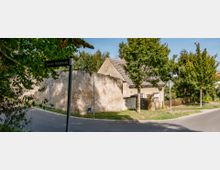 Straßenecke mit einem alten Wohnhaus mit Ziegeldach und angrenzender Steinmauer, umgeben von Bäumen. Ein Straßenschild mit der Aufschrift „Glindower Mühlenstraße“ ist im Vordergrund zu sehen.