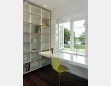 Ein modernes, schlichtes Arbeitszimmer mit einem weißen Wandregal, einem weißen Schreibtisch, einem grünen Stuhl und einem offenen Fenster mit Blick auf einen Garten. Dunkler Holzboden und minimalistische Dekoration runden den Raum ab.