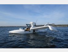 Ein kleines weißes Wasserflugzeug mit offenem Cockpit treibt auf einer ruhigen Wasseroberfläche, umgeben von einer weiten Landschaft und einem klaren Himmel.